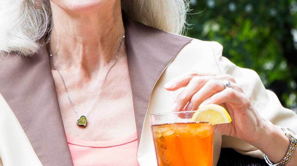 Close-up of an older woman wearing a jacket, pink t-shirt and green heart necklace while holding her hand over a glass of iced tea with a slice of lemon.