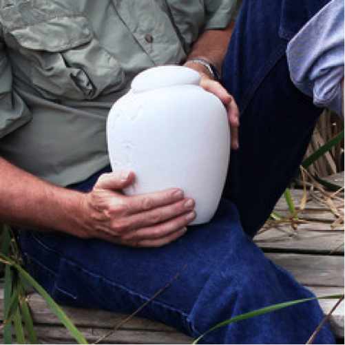 Close-up of the torso and hands of a man sitting outside holding a white clay cremation urn.